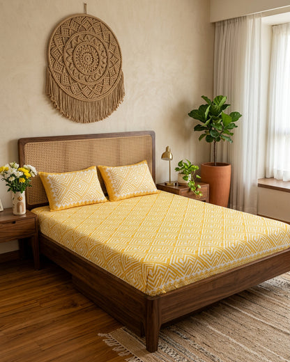 Aakriti Yellow bedsheet, marigold-yellow diamond-maze print, on a bed with a cane headboard with macramé