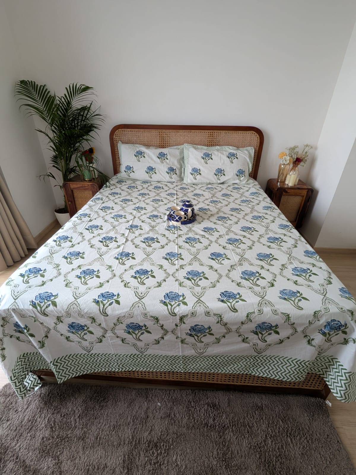 Bed with blue floral patterned bedsheet and pillows in a room with a plant and nightstands.
