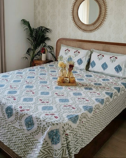 Bedroom with a bed covered in a patterned bedsheet, reversible pillows, and a small table with decor items.