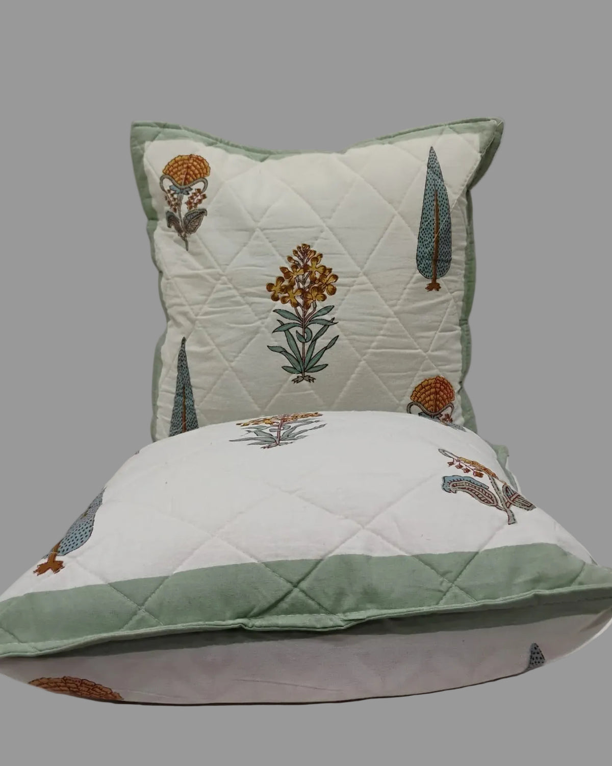Two Gulmohar green quilted cushion covers stacked to show the printed fronts and green trim