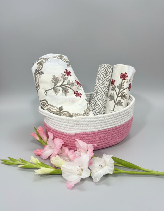 Cotton bedsheet floral patterns and 2 pillow covers, folded in a pink woven basket, surrounded by flowers.