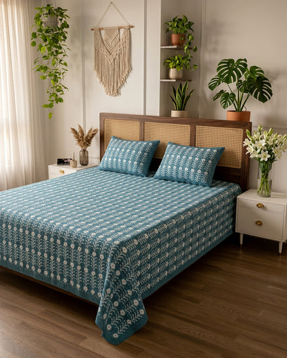 Harsingar Blue bedsheet, muted teal-blue ground with rows of white flowering stems, on a bed with a cane-panel headboard with plants
