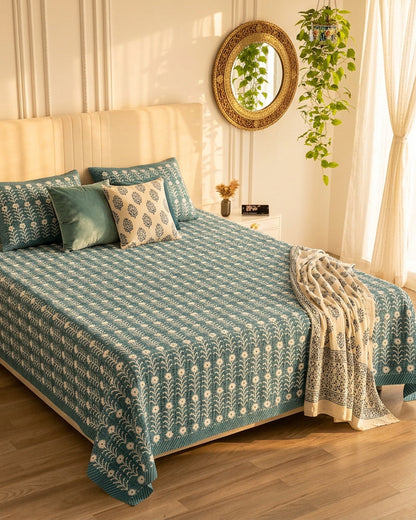 Harsingar Blue bedsheet, muted teal-blue ground with rows of white flowering stems, in a sunlit bedroom with a knit throw