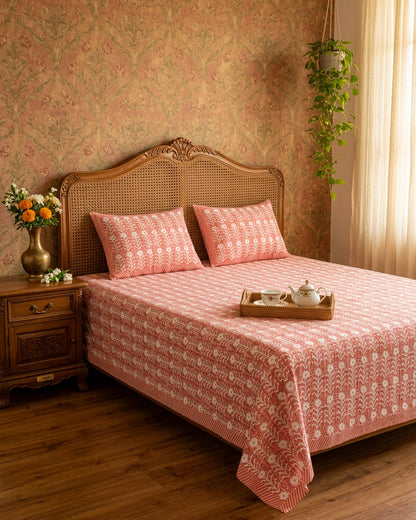Harsingar Pink bedsheet, coral-pink ground with rows of white flowering stems, on a bed with a cane headboard and a tea tray