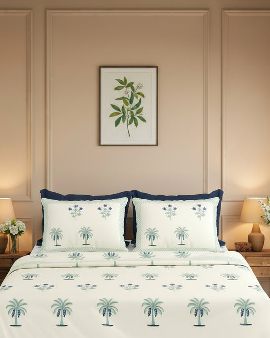 A neatly made bed in a styled bedroom setting, featuring a cream-colored duvet cover and matching pillowcases printed with blue palm trees and floral motifs. The bed is flanked by two wooden nightstands with lit table lamps, and a large arched wooden headboard is mounted on the wall behind.
