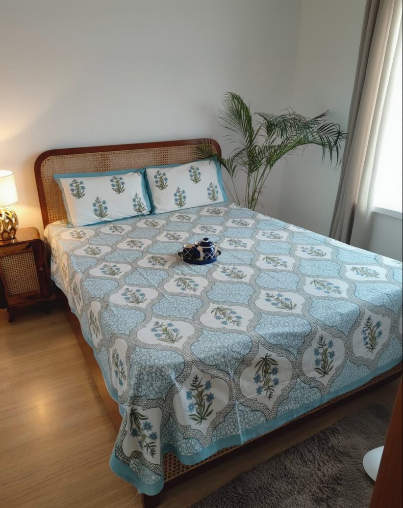 Bedroom with a bed covered in a blue patterned bedsheet and pillows, next to a nightstand with a lamp.