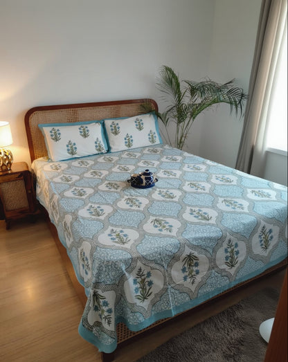 Bedroom with a bed covered in a blue patterned bedsheet and pillows, next to a nightstand with a lamp.