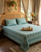 Sagar Blue bedsheet, teal geometric maze with a medallion-flower border, on a bed with a cane headboard and a tea tray