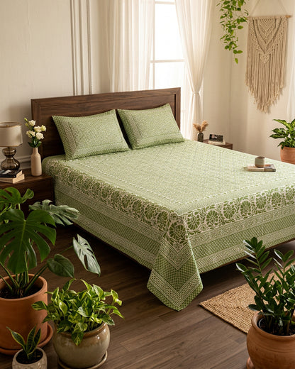 Sagar Green bedsheet, sage-green geometric maze with a medallion-flower border, on a bed with a dark wood headboard with potted plants