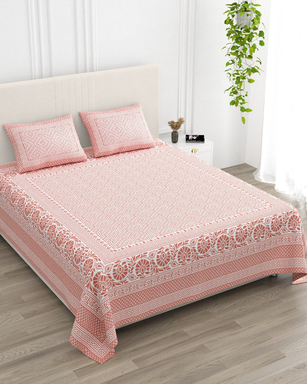 Sagar Pink bedsheet, coral-pink geometric maze with a medallion-flower border, in a studio shot against a cream headboard