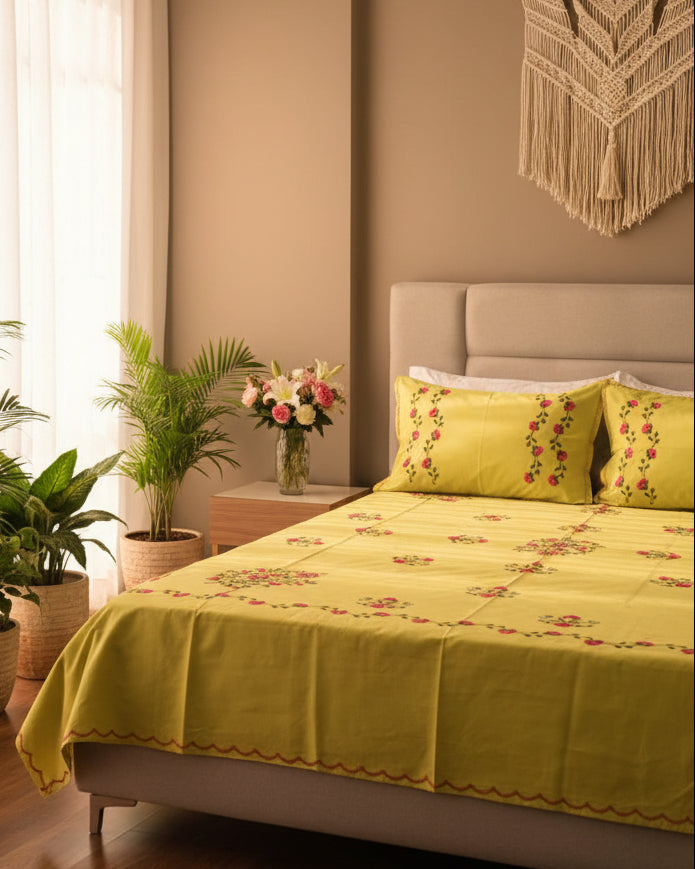 A bed dressed in a bright yellow-green bedsheet set with matching pillowcases, decorated with small pink floral motifs and green vine borders. The bed has a dark purple tufted headboard, with a bedside table and flowers visible in the background.