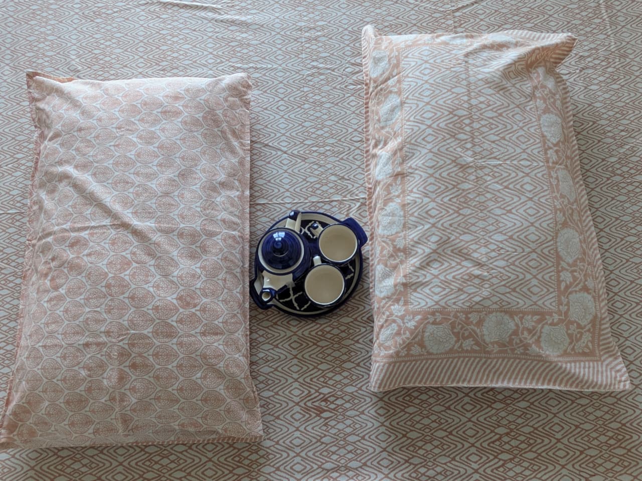 Two patterned pillows in peach on a bedsheet with a kettle and cup in between