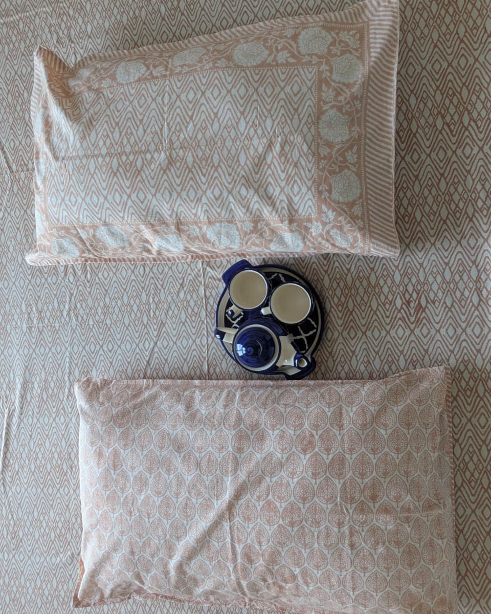 Two patterned pillows in peach on a bedsheet with a kettle and cup in between