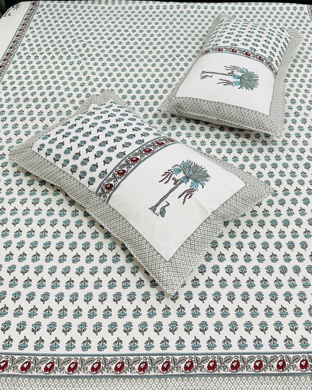 A white cotton bed sheet and matching pillowcases featuring a repeating blue-gray floral block print and a decorative border with red paisley accents. Two matching pillowcases are displayed on the bed.
