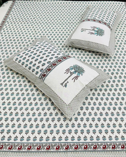 A white cotton bed sheet and matching pillowcases featuring a repeating blue-gray floral block print and a decorative border with red paisley accents. Two matching pillowcases are displayed on the bed.