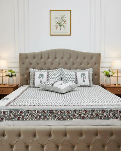 A white cotton bed sheet and matching pillowcases featuring a repeating blue-gray floral block print and a decorative border with red paisley accents. Two matching pillowcases are displayed on the bed.
