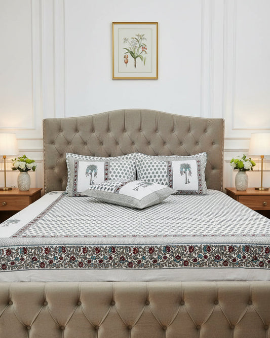A white cotton bed sheet and matching pillowcases featuring a repeating blue-gray floral block print and a decorative border with red paisley accents. Two matching pillowcases are displayed on the bed.