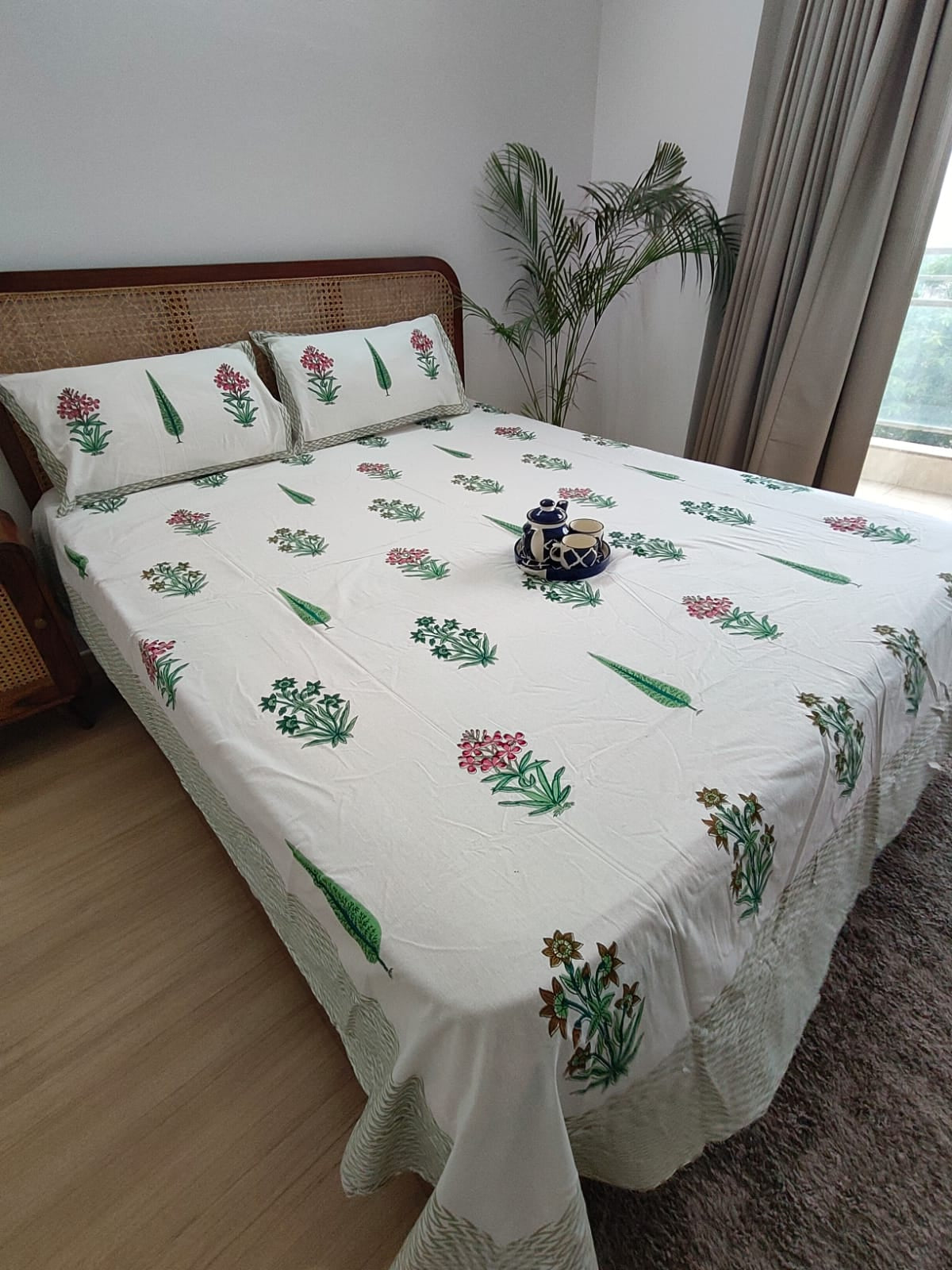 Bed with floral-patterned bedding in a room with a plant and window.
